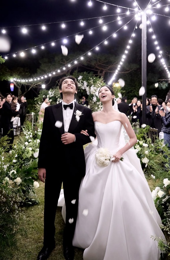 屋外の結婚式で、黒いタキシード姿のソン・ジホと白いドレス姿のキム・ソリが腕を組んで笑顔を見せる