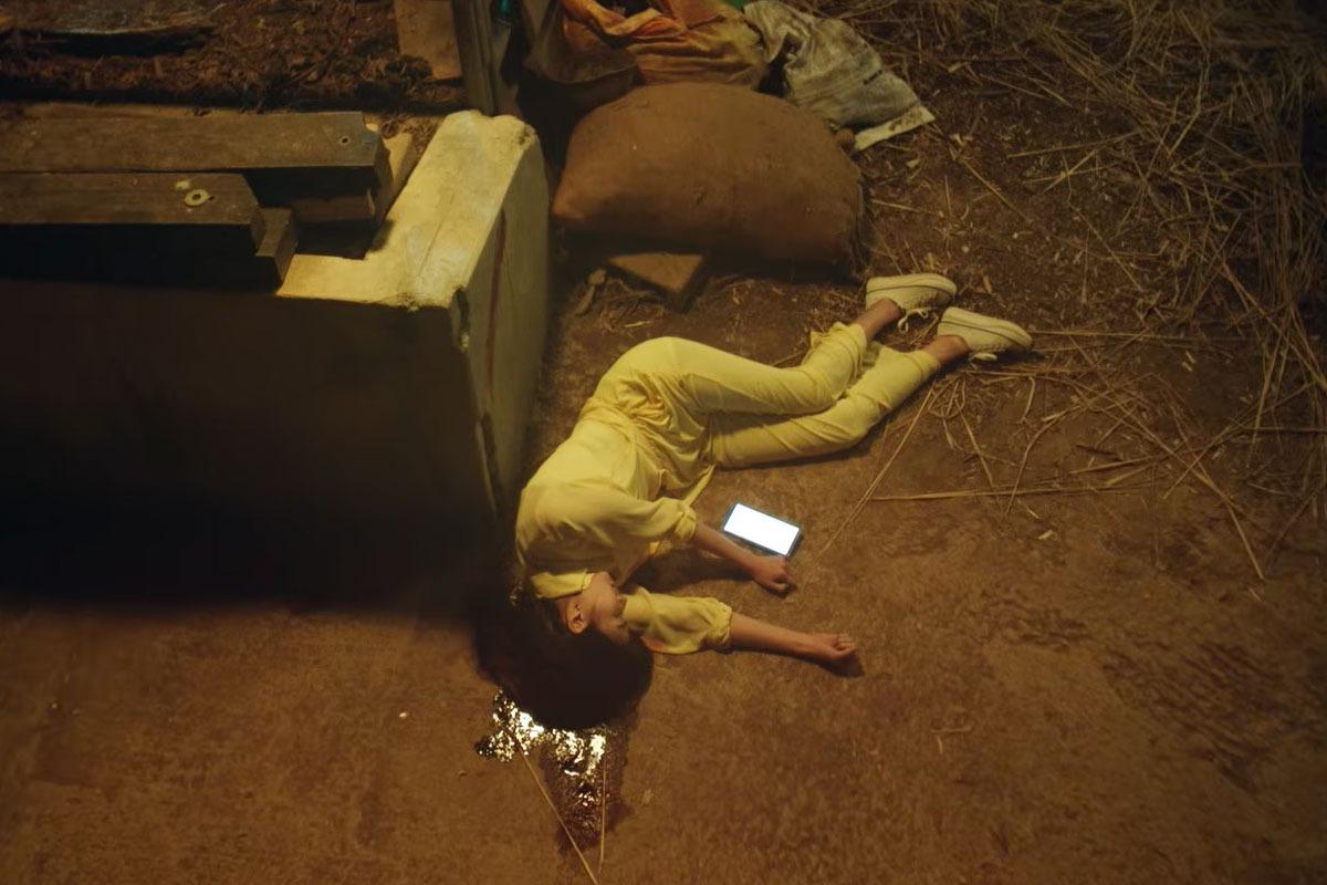 暗い場所で黄色い服の人物が地面に横たわり、そばに光るスマートフォンが見える『夏休み』の場面写真