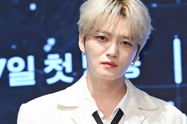 濃いブロンドの髪で白いジャケット姿のキム・ジェジュンが会見場で正面を向く
