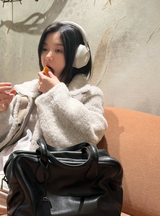 白いボアジャケットとヘッドホンを身に着け、ミカンを食べるキム・ユジョン
