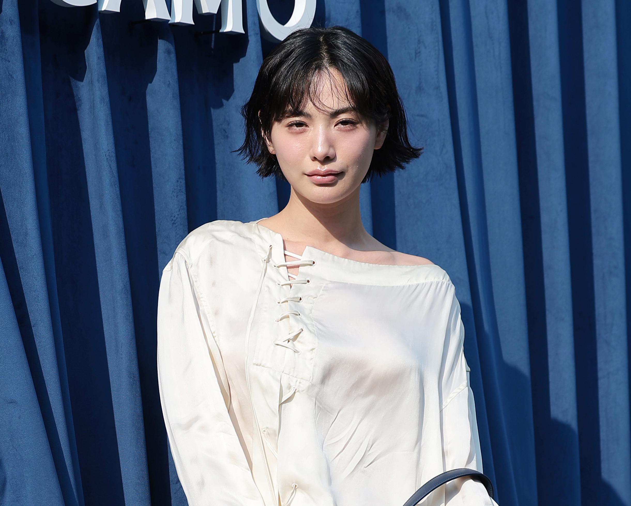 青い背景の前で白いブラウス姿でポーズをとるナナ